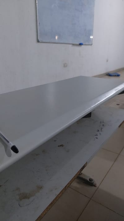 The image shows an interior scene, likely a classroom or office, in New Nyanya, Nigeria. A light grey, rectangular desk surface dominates the foreground, angled towards the viewer. The desk appears to be made of a smooth, possibly laminate material. A black pen with a silver clip rests on the left edge of the desk. Beneath this desk, another similar desk surface is visible, showing signs of wear and possibly some faint writing or smudges. The background features a white wall with a light blue rectangular whiteboard mounted on it. Below the whiteboard, a small blue object, possibly an eraser, is visible. The floor appears to be tiled in a light brown or beige color. The overall impression is of a functional, albeit somewhat aged, workspace.