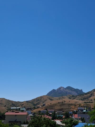 The image shows a panoramic view of a hillside village under a clear blue sky. The foreground is dominated by lush green trees, partially obscuring several buildings. The most prominent structure in the foreground is a two-story house with a red metal roof and a pale yellow facade. Behind this house, other buildings with red, white, and grey roofs are visible, nestled amongst the greenery.
The middle ground of the image transitions into a dry, hilly landscape dotted with more houses and structures. The hills are covered in dry grass and scrub, with terraced fields visible on some slopes. A large, white, elongated building, possibly a warehouse or industrial structure, stands out on the left side of the middle ground. Further into the scene, a tall, slender tower-like structure is visible on one of the hills.
In the background, a majestic mountain range rises, its rocky peaks casting shadows. The mountains are shades of grey and brown, with some vegetation clinging to their lower slopes. The overall impression is a sun-drenched, rural landscape. The scene appears to be situated within the Щебетовский поселковый совет in Ukraine. There are no people visible in the image.