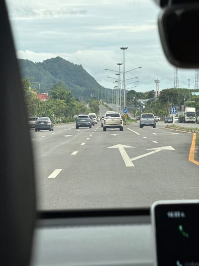 The image captures a view from inside a vehicle traveling on a multi-lane road in Ban Ta Khun, Thailand. The road is paved with asphalt and marked with white lane lines and directional arrows. Several vehicles, including sedans, pickup trucks, and a large truck, are visible on the road, moving in the same direction. The surroundings include lush green trees lining the roadside, tall streetlights, and distant mountains under a cloudy sky. A water tower and telecommunications towers are also present in the background. The interior of the vehicle is partially visible, showing a car dashboard with a screen displaying some information.