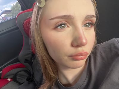 A close-up, low-angle shot features a young woman in the foreground, her face turned to the right. She has light brown hair styled down, with two light-colored hair clips visible. Her makeup includes pink blush on her cheeks, mascara, and lip gloss. She is wearing a grey t-shirt. In the background, out of focus, is a car seat with red and grey fabric. The scene appears to be indoors or in a shaded area, as indicated by the dark background to the right of the woman. The context of Novaya Otradovka, Russia, is not visually supported by the image content.