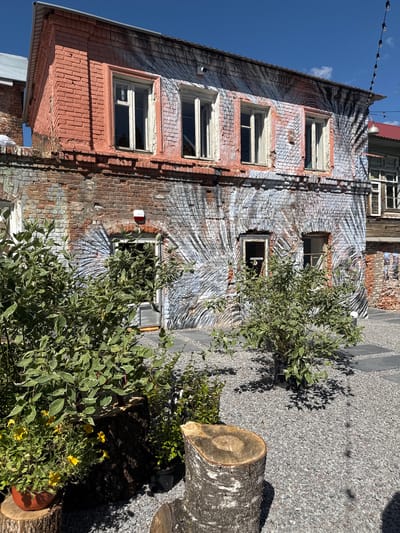The image depicts the exterior of a weathered, two-story brick building in Yelets, Russia, with a unique mural art style covering its facade. The lower part of the building displays exposed brickwork in various shades of red and brown, showing signs of age and wear. The upper portion of the building is painted with a stylized mural featuring broad, sweeping strokes of gray and white, resembling palm fronds or a sunburst pattern, interspersed with areas of pinkish-red paint.
Several windows are visible on both stories. The upper windows are rectangular with white frames, some appearing to be boarded up or in disrepair. The lower windows also have white frames, and one of them leads to a doorway with a gray door. A red fire alarm or emergency light is mounted on the wall near the lower doorway.
In the foreground, a landscaped area is covered with gray gravel. Several large tree stumps are placed as decorative elements. Lush green plants, including a large shrub with variegated leaves and yellow flowers in a pot, are arranged around the stumps. The lighting suggests a bright, sunny day, with clear blue skies overhead. In the background, another building with a red roof is partially visible, along with overhead string lights. The overall impression is of an urban courtyard that has been creatively revitalized with art and landscaping.