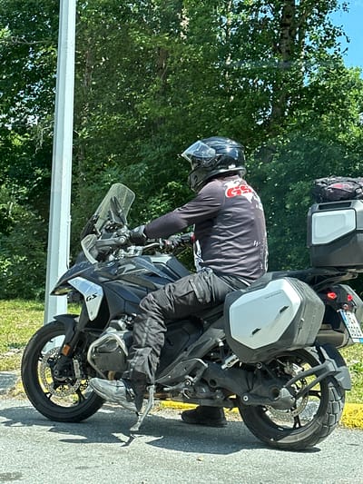 A man wearing a black helmet and a black long-sleeved shirt with red and white graphics is seated on a black BMW R 1250 GS motorcycle. The motorcycle is equipped with silver panniers on both sides and a black top case at the rear, which holds a red and black bag. The man is wearing black motorcycle pants and black boots. He is positioned on the right side of the motorcycle, with his left foot on the kickstand, and appears to be parked or waiting. The motorcycle is situated on an asphalt road with a yellow line marking the edge. In the background, there is a white utility pole and lush green trees. The scene is likely located outdoors, under daylight, with a clear sky visible through the foliage. The license plate on the rear of the motorcycle reads "TE 3629". The specific location within Crkvine, Montenegro cannot be determined from the visual information alone.