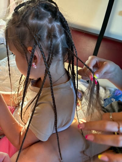 A person's hands are braiding the hair of a young child, who is facing away from the camera. The child has dark hair styled into many thin braids. The person braiding the hair has bright pink and yellow nail polish. The scene appears to be indoors, possibly on a bed, with a red headboard and white bedding visible. The lighting suggests a warm, natural light source. The location of Rossosh, Russia is not visible in the image.