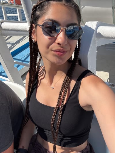 A young woman with dark, braided hair and sunglasses is taking a selfie outdoors. She is wearing a black tank top and a silver necklace with a small pendant. She is sitting on a white bench, with a blue railing and the sea visible in the background. The scene appears to be on a ferry or a boat, given the presence of railings and the open water.