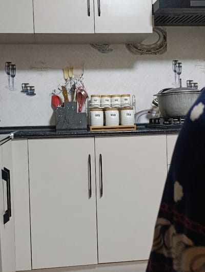 A kitchen scene shows white cabinets above and below a black countertop. The upper cabinets have silver handles. On the countertop are cooking utensils in a dark holder, a spice rack with jars labeled "Salt," "Flour," "Spice," "Coffee," "Sugar," and "Tea," and a large pot on a stove. Part of a blue and brown patterned garment is visible on the right side of the image, partially obscuring the view. The backsplash has a decorative design with wine glasses and other motifs.