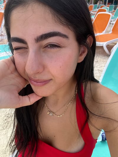 A close-up image features a female individual located in the Berry Islands, Bahamas. The subject has long, dark hair and fair skin, with prominent dark eyebrows. She is observed winking with her right eye closed and her left eye open, displaying a slight smile. Her right hand, featuring a ring on the ring finger, is positioned with fingers resting under her chin. She is wearing a red bikini top and two gold-toned necklaces, one of which includes a small charm cluster. The background reveals a sunny beach environment with light-colored sand. Multiple rows of empty beach chairs, alternately colored orange and light blue/teal, are visible in the mid-ground. A clear plastic water bottle with a blue cap is partially visible on a light blue/teal chair to the right of the subject.