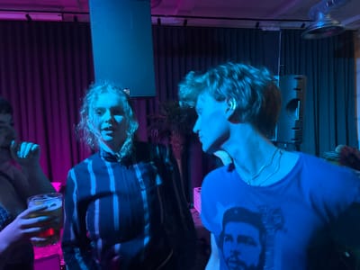 The image depicts three individuals in a vibrant, dimly lit indoor setting, likely a bar or music venue, illuminated by strong blue and pink/purple artificial lights.
In the left-center of the frame, a young woman with curly blonde hair and a lip piercing is shown from the chest up, wearing a dark striped long-sleeve top. Her mouth is slightly open, suggesting she might be talking, singing, or reacting to something. To her right, and slightly closer to the foreground, a young person with short, wavy light hair is seen in profile, looking towards the left. They are wearing a blue t-shirt prominently featuring a black and white graphic of **Che Guevara's** face. This individual also sports an ear cuff and a delicate necklace. Partially visible on the far left, a third person holds up a glass containing an amber liquid, likely beer, with their hand.
The background is dominated by dark, heavy curtains, possibly indicating a stage or performance area. Two large black speakers are positioned on either side of the frame, with their mesh grilles visible. A potted plant, resembling a palm, is situated between the two main subjects, and a pink plastic crate can be seen behind the person in the blue t-shirt. Overhead, a metallic air duct or vent is visible on the ceiling in the upper right corner.
The scene conveys a social event or gathering, likely an evening or nighttime affair due to the mood lighting. The presence of speakers and drinks suggests music or entertainment is central to the event. The emotions of the individuals are not clearly discernible, but the overall atmosphere appears relaxed and social. No explicit location cues link the image to Dundee, United Kingdom, but it fits the description of a typical indoor entertainment space.