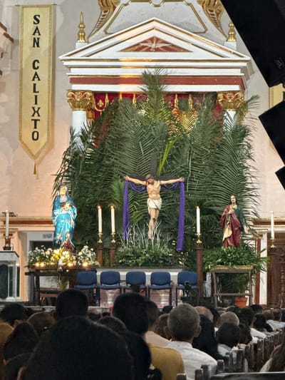 The image depicts a church interior in Timaná, Colombia, with religious statues and decorations visible behind a seated audience. The altar features a depiction of Jesus on the cross, flanked by statues of other religious figures amidst green foliage. A banner reading "SAN CALIXTO" is hung to the left of the altar. Rows of chairs are occupied by people, mostly with dark hair, facing the altar. The scene is captured with an informal, candid style, seemingly from within the audience.