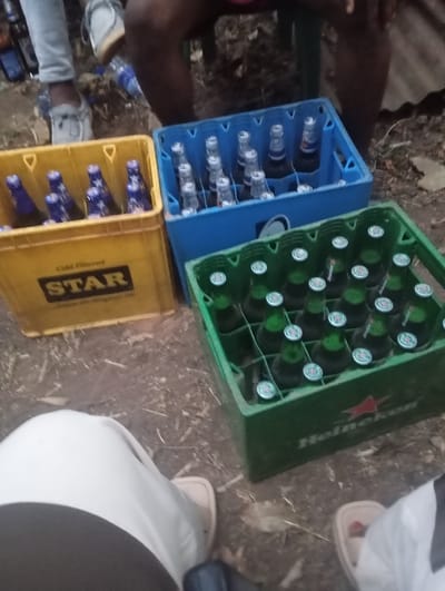 Three crates of beer sit on the ground outdoors. A yellow crate marked "STAR" holds dark bottles with blue labels. A blue crate beside it contains brown bottles with silver caps. The green crate closest to the camera has the Heineken logo and is full of green bottles with silver caps. Parts of people's legs and feet are visible around the crates. The ground is dirt with scattered twigs and leaves.
