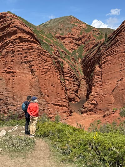 The image captures a vibrant natural landscape, specifically in the Jeti-Ögüz region of Kyrgyzstan, characterized by its striking red rock formations. Two individuals are the main subjects, viewed from behind, standing on a dirt path at the edge of a vantage point. They appear to be observing the vast canyon and towering cliffs before them.
One person, wearing a bright orange hooded jacket, tan trousers, and white shoes, has visible grey hair and carries a blue backpack. The other person is dressed in a dark blue or black jacket and dark trousers, also with a backpack, possibly blue or teal in color.
The scene is dominated by massive red sandstone cliffs that rise steeply, showcasing pronounced horizontal stratification and intricate erosion patterns that have carved out deep grooves and ledges. Green vegetation, including shrubs, bushes, and grasses, clings to the slopes and grows along the pathway, with small clusters of yellow wildflowers visible in the foreground. Below the cliffs, a lighter dirt path or canyon floor can be seen winding through the red rock.
The sky is a clear, brilliant blue with scattered, fluffy white clouds, suggesting a sunny and pleasant day, likely late morning or early afternoon, ideal for hiking and sightseeing. There is no visible text in the image. The overall impression is one of awe-inspiring natural beauty and exploration.