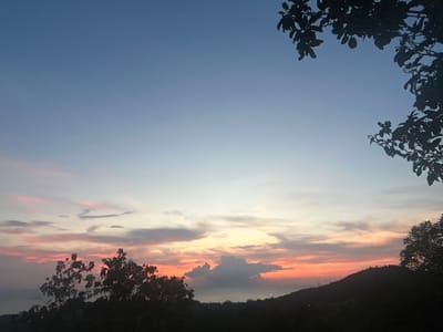 The image depicts a sunset in Baan Hin Kong, Thailand. The sky transitions from blue at the top to pink and orange hues near the horizon. There are clouds, with a prominent gray one centrally positioned. Silhouetted trees and a hill line the bottom of the image. Tree branches and leaves are visible in the top right corner.