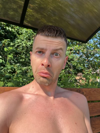 An individual with short brown hair and a light complexion is positioned in the foreground, looking directly at the camera with a surprised or confused expression. They are shirtless and appear to be outdoors, seated on a wooden bench. Behind them, lush green foliage creates a backdrop, suggesting a natural or garden setting. The upper portion of the frame is partially obscured by a translucent, corrugated roof structure, possibly part of a gazebo or shelter. The lighting indicates it is daytime, with sunlight casting shadows. The individual's skin shows some redness, indicative of sun exposure. No specific landmarks or buildings are visible that would identify the location as Navahrudak, Belarus.