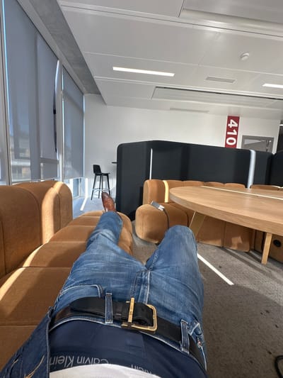 The image depicts a first-person, low-angle view from a reclining position within a modern office or lounge environment located in Clichy, France. The foreground shows the lower torso and legs of an individual. The individual wears blue denim jeans, a black leather belt with a rectangular gold-colored buckle, and a visible dark blue waistband with "Calvin Klein" branding. The right leg is extended, terminating in a polished brown leather dress shoe.

The individual is reclining on a light brown, segmented, upholstered modular sofa that extends to the left and into the midground. To the right, a round, light wood table with a central base is positioned next to another segment of the modular sofa. A small, metallic gray electronic device with an attached cable rests on this sofa segment. Behind the table and sofas, a dark gray or black high-backed booth or privacy partition is visible.

Architectural features include large windows along the left side of the frame, partially covered by white translucent roller blinds, allowing direct sunlight to enter and cast strong shadows. The room has white walls and a white suspended ceiling with integrated rectangular LED light fixtures and air conditioning vents. The floor is covered with light gray carpeting. On a white wall in the background, a red vertical sign with the white numbers "410" is prominently displayed, indicating a room or zone number. A single black high-stool chair stands near the windows in the background.