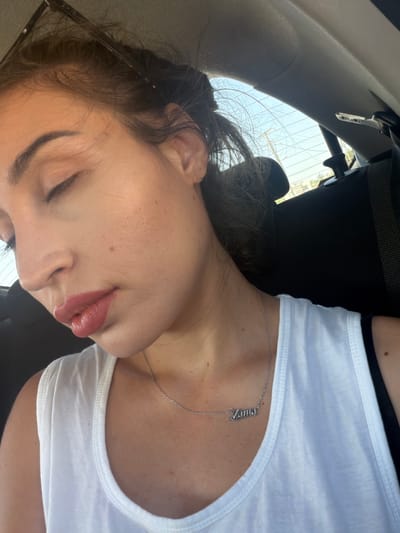 A close-up, over-the-shoulder shot captures a young woman with her eyes closed, appearing to be asleep or resting in the back seat of a car. She is wearing a white tank top and a silver necklace with a pendant that reads "Mama." The setting appears to be within a vehicle, suggested by the car's interior elements like a headrest and seatbelt visible in the background. The lighting indicates it is daytime, with sunlight filtering in. The woman has on makeup, including lipstick. The car is parked or moving slowly on a road that shows a glimpse of trees and power lines through the rear window. The location is identified as Dromolaxia - Meneou Municipality, Cyprus.