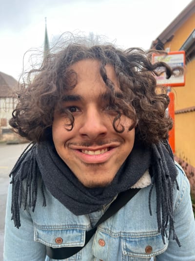 A young adult male is depicted in a close-up, wide-angle portrait against an outdoor urban background in Meistratzheim, France. He possesses dark, voluminous curly hair, a thin mustache, and a goatee. The male is looking directly at the camera with a broad smile, revealing his teeth. He is dressed in a dark gray knitted scarf with fringed ends and a light blue denim jacket featuring a white faux-shearling collar. A dark strap from an unseen bag is visible across his chest. The background on the left features a portion of a traditional half-timbered building and the upper spire of a church, dark and slender, against an overcast sky. On the right, a building with an orange-yellow facade and a terracotta-tiled roof is visible, along with a partially obscured light-colored sign bearing blue text.