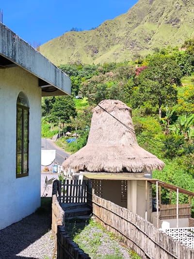 The photograph captures a rural scene in Sembalun Bumbung, Indonesia. The foreground is dominated by a white stucco building with a tall, arched window. Adjacent to the building is a wooden fence that runs along a stone path leading to a small structure with a thatched roof resembling a traditional Indonesian hat. The thatched roof structure has open sides with bamboo detailing and a raised floor. In the background, a lush green hillside rises steeply, covered in trees and vegetation. A road is visible winding through the trees, and a few small structures can be seen in the distance. The sky is bright blue, suggesting a clear day. Electrical wires are visible running across the scene, cutting through the natural landscape.