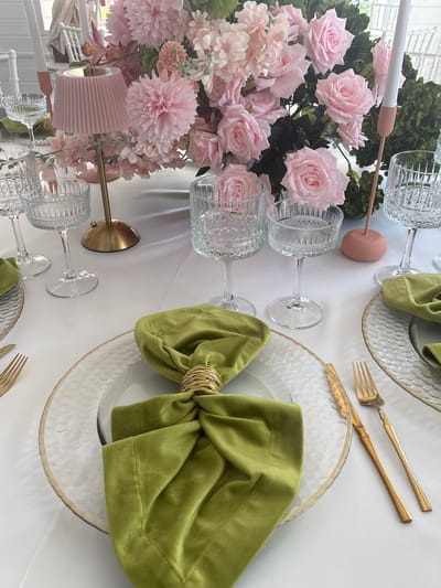A close-up shot shows a beautifully decorated table setting, likely for a formal event such as a wedding or upscale dinner party. Pink roses and other flowers, arranged in a bouquet, occupy the left side of the frame. Crystal glassware is interspersed throughout the floral arrangement.

To the right of the flowers are dinner plates, each adorned with a green velvet napkin secured with a gold napkin ring. Gold-colored forks and knives are positioned next to the plates. The plates appear to be made of textured glass with a gold trim.

The table is covered with a white tablecloth. The background is indistinct but suggests an indoor setting with natural light. The scene lacks human presence.
The location in the city of Юг, Russia cannot be determined from the provided image.