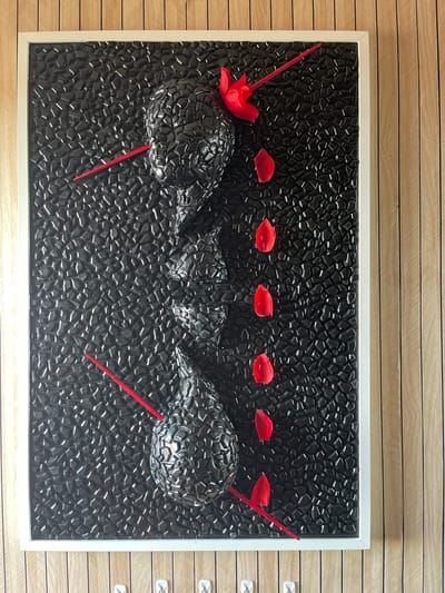 The image shows a framed artwork with a black, textured background composed of irregular, raised shapes. A sculpted, dark gray, abstract representation of a human profile is embedded in the background, facing left. Red elements are incorporated into the artwork, including thin, rod-like shapes piercing the profile and scattered petal-like shapes. The artwork is mounted on a wall with vertical wooden planks. There are no people visible in the image, and there is no discernible location in the city of Portsmouth, United Kingdom.