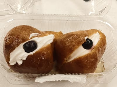 Two rum baba pastries are presented in a clear plastic container. Each pastry is golden brown and glazed, with a dollop of white cream and a dark berry on top. The container is slightly wet with condensation. The pastries are arranged side-by-side. The image does not contain any information about the city of Pozzuoli, Italy.