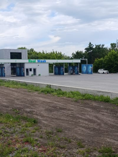 This is a wide shot of a car wash facility in Khorol, Ukraine, under a cloudy sky. The car wash is a modern, multi-bay structure with a prominent green sign that reads "Real Wash." To the left of the main structure, there is a section with vertical metallic siding. Several bays are visible, some with blue panels displaying a water droplet logo. In one bay, two individuals appear to be working on a vehicle. Further to the right, a white SUV is parked in a bay. The facility is situated on a paved lot, with a low concrete barrier separating it from a dirt area in the foreground. The surrounding area is lined with dense green trees, and the ground in the foreground is a mixture of dirt, small stones, and sparse vegetation.