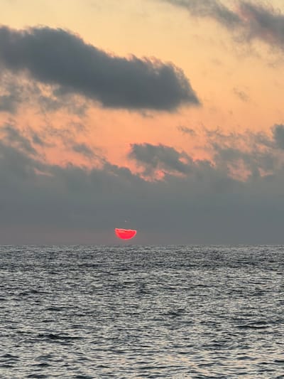 A vertically oriented image shows a striking contrast between sky and sea. The sky, on the left, is filled with dramatic clouds tinged with peach and grey. A solitary red parachute, with a figure dangling beneath it, punctuates the sky between the clouds and the rough, textured surface of the sea. The sea extends horizontally across the right of the image, marked by a repetitive wave pattern.
