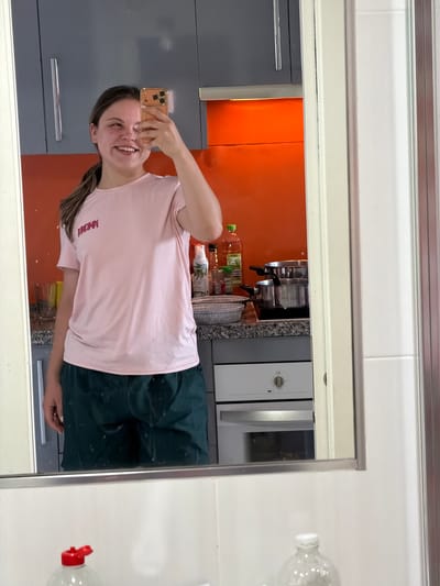 A young woman is taking a mirror selfie in a kitchen. The woman has brown hair and is wearing a pink t-shirt and dark green shorts. She is smiling and holding her phone up in her right hand.
The kitchen has grey and orange cabinets, a stainless steel oven, and tiled walls. There are bottles of oil and other cooking items visible on the counter.
The location is most likely Arroyo de la Miel-Benalmádena Costa, Spain, as specified in the request.