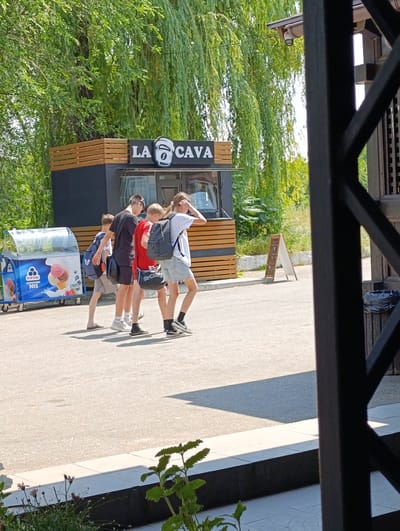 In Ladyzhyn, Ukraine, on a sunny day, a group of four young people walks past an ice cream cart and a food stand. The food stand, labeled "LA CAVA" with a logo of a coffee cup, is decorated with wooden slats and has a black exterior. To the left, a blue and white ice cream cart displays images of ice cream cones and tubs with the text "РУДД МОРОЗИВО №1". The people are dressed casually in t-shirts, shorts, and backpacks, suggesting a leisurely outing. The scene appears to be outdoors, possibly in a park or near a recreational area, with lush green trees providing shade. The lighting indicates it is daytime, likely mid-morning or afternoon. The atmosphere seems relaxed, with the individuals walking at a comfortable pace. A white A-frame sign with images of coffee cups is visible further in the background. The overall impression is a typical summer day in a public space.