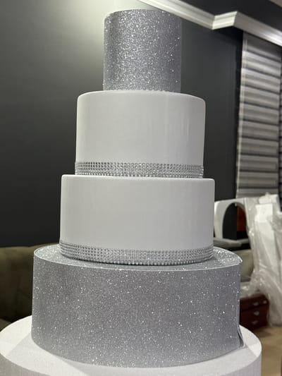 The image shows a four-tiered cake, likely a wedding cake, with a silver glitter top tier and silver glitter bottom tier. The two middle tiers are white and decorated with a silver band of rhinestones. The cake is placed on a white styrofoam base. In the background, a dark grey wall is visible, with a window covered by blinds on the right. There are also some out-of-focus objects in the background, including what appear to be plastic sheeting and furniture. The scene is indoors, suggesting a home or event venue. The location of Gölbaşı, Turkey is not discernible from the image itself.