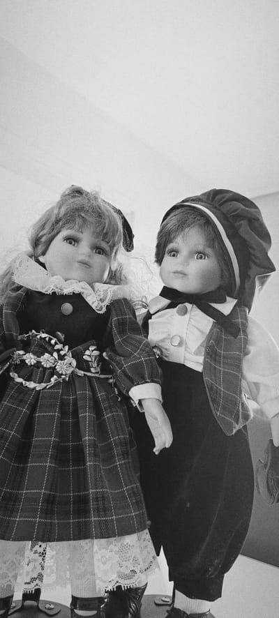 Two porcelain dolls are depicted from a low angle, with a white wall visible in the background. The doll on the left, presumed female, wears a plaid dress with a white lace collar and trim, and has blonde hair tied with a black ribbon. The doll on the right, presumed male, is dressed in a waistcoat, white shirt with a bow tie, and a hat, with dark hair. The dolls are positioned close together, facing forward. The scene is monochromatic. There is no information available to suggest this image is located in Winnenden, Germany.