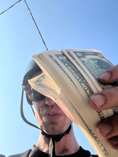 The image is a close-up, low-angle shot of a person holding a fan of US dollar bills in front of their face. The person is wearing sunglasses and a helmet with a chin strap. The background is a clear blue sky. The dollar bills appear to be two-dollar bills, with "TWO" visible on some of them. The person's lips are parted, and they are looking towards the camera. The lighting suggests it is daytime, possibly outdoors. There is no indication of the location being in the city of Mussolini, Italy, or any other specific city.