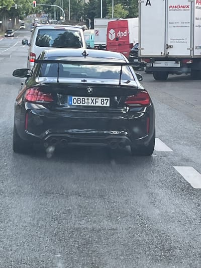 The image shows a black BMW M2 Competition with a large rear spoiler and quad exhaust pipes, parked in traffic on a city street. The license plate reads "OBB XF 87". In the background, there is a white box truck with "PHÖNIX" logo and "Gepäcktransport & Umzüge" written on it, which is a company based in Aschaffenburg, Germany, as indicated by the address "Reisingerstraße 17 (63741) Aschaffenburg" visible on the truck. There are other vehicles, traffic lights, and trees visible in the background, suggesting an urban environment. The overall scene depicts a moment captured in a street in Aschaffenburg, Germany, with the BMW being the central focus.