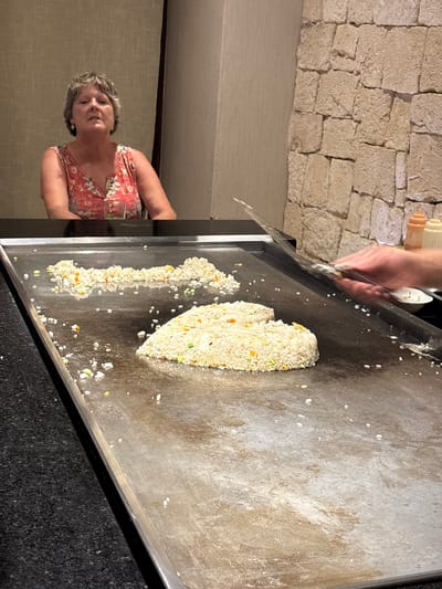 The image depicts a hibachi grill in Punta Sam, Mexico. A woman with short, graying hair and a floral print dress is seated at the grill. Two mounds of fried rice, one larger than the other, are on the hot surface of the grill. A hand with a spatula is visible on the right side of the image, in the process of cooking. The background features a stone wall and a textured wall. Condiment bottles are visible on a shelf behind the grill.