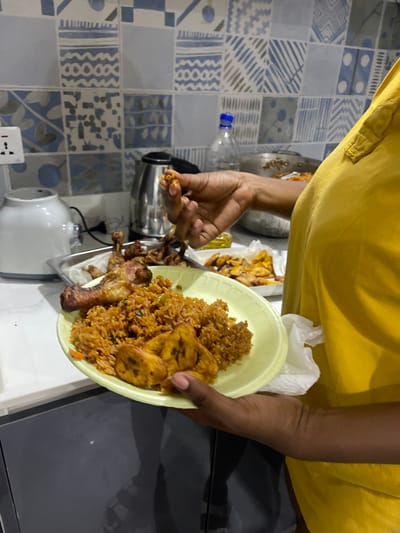 An overhead shot, rotated 90 degrees clockwise, captures a person holding a plate of food in a domestic kitchen setting in Adenta Municipality, Ghana. The person, wearing a yellow top, has dark skin and their hands are prominent in the frame. In their left hand, they hold a light-colored, possibly disposable, plate filled with a generous portion of Jollof rice, several slices of fried plantain, and a fried chicken drumstick. Their right hand holds a small, bite-sized piece of what appears to be fried food, possibly chicken or plantain.
In the background, more fried plantains and chicken pieces are visible on a tray lined with white paper towels. The kitchen counter beneath the food and appliances is a mix of white and grey. Various kitchen items are present, including a clear plastic water bottle with a blue cap, a silver blender, and a white kettle. An electrical outlet with a single socket is also visible on the lower left. The floor or wall features a distinctive pattern of blue, grey, and white tiles, some with intricate geometric designs. The scene depicts a moment of food preparation or serving, highlighting a meal common in West African cuisine. There is no visible text.