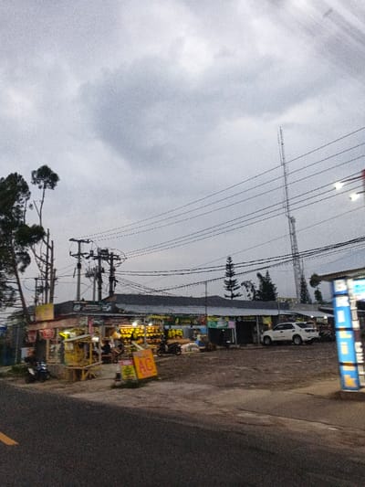 An overcast outdoor scene depicts a commercial strip in Phước Hòa, Vietnam, observed from a low angle across an asphalt road. The road, with a partial yellow line visible on the left, transitions into an unpaved dirt and gravel area that serves as a parking lot. In the midground, a row of single-story commercial buildings with corrugated metal roofs runs horizontally. The leftmost building, a shop, displays numerous items, possibly produce, illuminated by bright yellow lights, with indistinct human figures present nearby and a smaller, makeshift wooden stall situated in front. Further to the right along the strip, additional storefronts are visible, one bearing the text "ĐỨC QUANG".
Vehicles in the scene include a dark motorcycle parked near the leftmost shop, another motorcycle further right, and a white SUV (resembling a Toyota Fortuner) parked in the dirt lot facing left. A bright yellow A-frame sign with red "AC" text stands on the left side of the dirt area. On the far right, a tall, vertical blue sign with white text is partially visible. The upper portion of the image features a heavy, overcast sky. Numerous black electrical wires crisscross the frame, supported by multiple utility poles with complex wiring. In the background, a tall, slender metal communication tower rises prominently on the right. Trees are visible on the far left and sparsely behind the buildings.