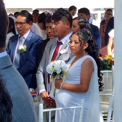 The image depicts a wedding ceremony, likely taking place in Cerro Colorado, Peru. A bride in a white dress is looking at the groom who is wearing a gray suit with a red tie and holding her hand. She is holding a white bouquet of flowers. They appear to be seated, perhaps on a white bench. Other attendees are visible in the background, dressed in suits and dresses, seated in rows. Some attendees are wearing sunglasses. The general setting suggests an outdoor venue, possibly with chairs set up for the guests.