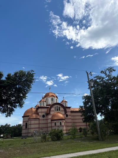 The image shows a large, domed building that appears to be a church, constructed with alternating bands of light and dark brick, and featuring multiple copper domes topped with crosses. The building is set against a bright blue sky with scattered white clouds. In the foreground, there is a patch of green grass and some scattered bushes. To the right of the building, a utility pole with electrical equipment stands tall, with power lines running horizontally across the frame. Trees are visible on both the left and right sides of the image, framing the view of the church. The overall scene depicts an exterior view of the church on a sunny day, with elements of urban infrastructure present.