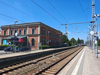 This media file depicts a sunny day at the Memmingen train station in Germany. The scene is dominated by the train station building, a red brick structure with multiple windows and an arched entrance. In the foreground, two sets of train tracks stretch into the distance, with gravel ballast and concrete sleepers visible. A train is seen approaching on the right track.
Various objects are present, including lampposts, overhead power lines for the trains, and signage. A prominent blue sign reads "Memmingen," indicating the location. Other signs include parking information ("P" with prices) and a platform number "1". A large clock on a lamppost near the station building shows the time. There are no people clearly visible in the image. The weather appears to be clear and sunny, with a bright blue sky. The overall impression is one of a functional and well-maintained European train station on a pleasant day.