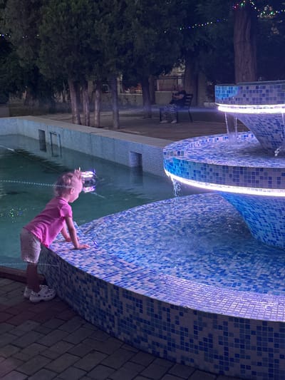 A young girl with blonde hair in a pink shirt and patterned shorts leans over a tiled fountain. The fountain is illuminated by blue and white lights and has water cascading down its levels. The girl's hands are on the edge of the fountain, and her body is angled towards the water. She appears to be looking down into the water. In the background, there are trees and what looks like a building. The scene appears to be outdoors at night, with string lights visible in the trees. The overall mood is peaceful and playful, with the girl enjoying the water feature. The location is identified as Bilhorod-Dnistrovskyi, Ukraine.