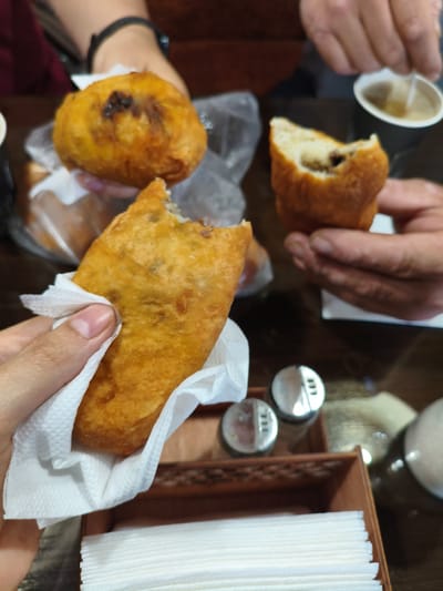 A close-up, overhead view shows hands holding and interacting with fried pastries on a wooden table. In the foreground, a hand is holding a golden-brown fried pastry wrapped in a white napkin. To the left of this pastry, another hand is holding a similar, whole pastry. In the middle ground, a partially eaten pastry with a dark filling is held by a person's hand. In the background, another whole pastry is visible. Two salt and pepper shakers sit in a wooden holder on the table, along with a stack of white napkins. A white paper cup with a light-colored liquid is also present. The scene appears to be indoors, likely in a casual dining setting. The context of Chernorech'e, Russia, suggests this could be a local bakery or cafe.