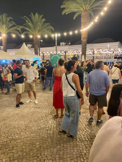 The image captures a lively night scene at an outdoor event in Tavira, Portugal, likely a festival or market, given the string lights and festive decorations overhead. The ground is paved with traditional cobblestones. In the foreground, several people are mingling. Two men are engaged in conversation, one in a dark t-shirt and beige shorts, holding a drink, and the other in a white button-down shirt and beige shorts. Behind them, a woman in a red dress and another in a white top and light blue jeans are visible. Further into the crowd, a man in a blue t-shirt and dark shorts is walking away from the camera, and other individuals are scattered throughout the scene, some appearing to be enjoying food or drinks at stalls. Tall palm trees punctuate the background, their fronds silhouetted against the night sky, with illuminated strings of lights draped between them. White tents, possibly for vendors, are set up in the middle ground. The overall atmosphere is one of social gathering and celebration.