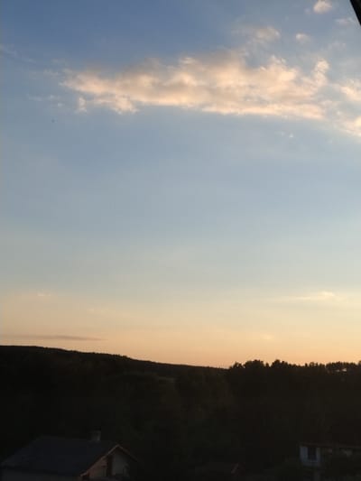 The image shows a rural landscape near Dobev, Czechia, at dusk. A dark, silhouetted forest stretches across the horizon, forming a low ridge. In the foreground, the roof of a building is visible on the left, and another building's corner is seen on the right. The sky is a gradient of soft blues and oranges, with scattered, pale clouds. Two small dark specks, possibly birds, are visible in the sky.