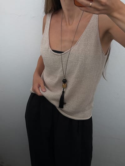 A person is modeling a sleeveless, light-colored knit top and black pants. The top has a delicate weave and a scoop neckline. A necklace with a black bead, a gold-toned woven bead, and a black tassel hangs from the neck. The person's left hand is visible, wearing a thin gold ring, and is holding what appears to be the edge of sunglasses. The background is a textured, off-white wall. The image is shot from a high angle and is oriented with the person's head to the left. The lighting is soft, suggesting an indoor setting with diffused light.