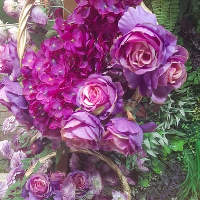 Two wicker baskets are filled with an arrangement of artificial purple and pink flowers, including roses and hydrangeas, and are placed in front of a backdrop of artificial greenery, resembling ferns and other plants. The scene is indoors, possibly a market stall or decorative display, and it is not possible to determine the specific location within the city of Sosnovka, Kyrgyzstan. There are no people or actions depicted in the image. The lighting is even, and the focus is sharp, allowing for clear detail of the floral arrangement.