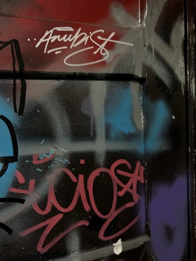 The image depicts a close-up view of graffiti on a rough, possibly metallic surface. The background is predominantly black with patches of blue, red, and purple paint. Overlaid on this backdrop are several layers of graffiti art. On the left side, there's a white, stylized signature-like design. To the right, there is what seems to be a pink, free-hand style text or symbol. The surface appears worn, possibly reflecting an outdoor setting. The location is Rio Grande, Argentina, but the graffiti is not indicative of a particular area, so more context would be needed to determine the specific neighborhood or building. There is no indication of people, interaction, or environment in this image.