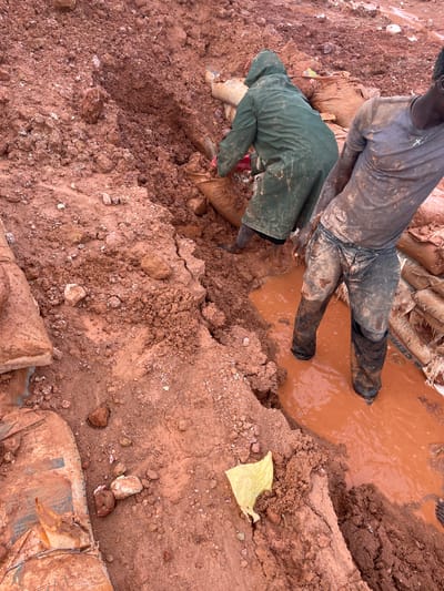 The image shows two people working in a muddy, reddish-brown environment, likely a construction or mining site. One individual, wearing a dark t-shirt and dark pants with tall black boots, is standing in a pool of muddy water. Their pants appear to be partially submerged. The other person is bent over, wearing a dark green rain jacket and also appears to be wearing boots. They are digging or working with materials in the mud.

The ground is uneven and covered in loose soil, rocks, and dirt. Piles of what look like sandbags are visible in the background, some with visible text. One sandbag has "CEMEN" clearly printed on it, suggesting it is a cement bag. A piece of bright yellow plastic is also lying on the ground in the foreground. The overall scene suggests a challenging and dirty work environment, possibly due to recent rain or ongoing excavation. The lighting indicates it is daytime. The location context is Rayfield, Nigeria.