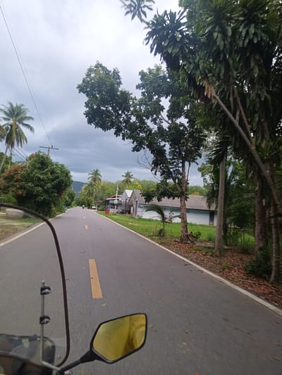 A gray asphalt road with a yellow center line stretches into the distance under an overcast sky. The road is flanked by lush vegetation including coconut palms and various trees. Buildings are visible along the roadside, including structures with gray roofs. The foreground shows the windscreen and side mirror of a motorcycle, implying the image was taken from the perspective of a person riding the motorcycle. The location is identified as Tambon Pak Phraek, Thailand. A distant figure on a motorcycle can be seen on the road.