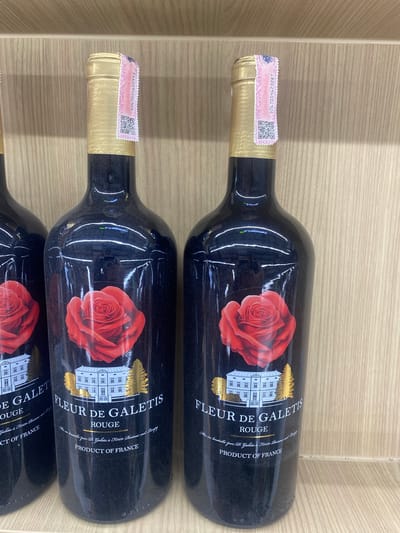 Two dark-colored wine bottles are prominently displayed, lying horizontally on a light wooden surface, possibly a shelf or crate. A third bottle is partially visible at the bottom edge of the frame. Each bottle is made of dark, almost black, glass and features a gold foil capsule covering the cork and neck.

The main label on each bottle is black with a striking red rose illustration on the left side. To the right of the rose, a white, château-like building with multiple windows is depicted. The primary text on the label reads "FLEUR DE GALETIS" in a stylized font, followed by "ROUGE". Below this, smaller text states "Mis en bouteille par D. Galetis à F11170 Raissac sur Lampy" and "PRODUCT OF FRANCE", indicating the wine's origin.

Around the neck of each bottle, a pink and white security or tax stamp is affixed over the gold foil. These stamps feature numerical codes and QR codes. The visible numerical codes are "0267054622993" on the top bottle and "0267054623287" on the middle bottle. The bottles appear to be new and some have minor reflections or dust on their surfaces. The setting is indoors, with the wooden background suggesting a retail display or storage area in Moo 1, Thailand. No human activity is depicted.