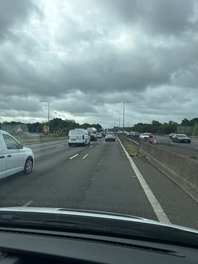 The image depicts a traffic jam or slow-moving traffic on a multi-lane highway, viewed from inside a vehicle. The perspective is forward and slightly to the right, with a portion of the originating vehicle visible on the right side of the frame.

Numerous vehicles, including cars and vans of various makes and colors, are present across several lanes. Most appear to be stationary or inching forward slowly. A white van is prominently featured in the lower right, indicating it's the vehicle from which the photograph was taken. Further down the road, another white van displays a partially visible UK license plate "Y270 K...".

The highway features a dark asphalt surface with clear white road markings, including a solid white line bordering the hard shoulder and dashed white lines separating lanes. A concrete barrier runs along the right side of the road, separating it from a treelined embankment. Tall streetlights or gantry structures are spaced along the highway.

The sky is heavily overcast with dense, grey clouds, indicating a dull or potentially rainy day. Visible road signs include a yellow diamond-shaped warning sign with a 'speed bumps' or 'uneven road' symbol in the middle distance, which is characteristic of UK road infrastructure. Another blue rectangular sign is visible further along but its text is illegible.

The setting is a busy motorway, consistent with the given location of Denham, United Kingdom, suggesting a typical traffic congestion event. No individuals are clearly visible.