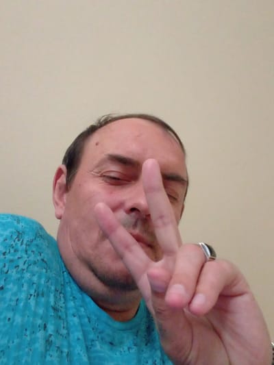 A man with light skin, dark hair, and stubble is pictured from the chest up, holding two fingers up in a peace sign. He is wearing a bright turquoise t-shirt with a dark, speckled pattern. The background is a plain, light beige wall. The man has a ring on his right ring finger. The image is taken from a low angle. The specific location in Kajaani, Finland cannot be determined from the provided image.