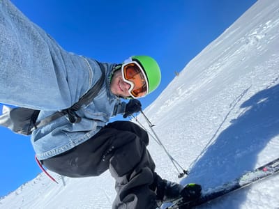 This image depicts a single individual taking an upward-angled selfie on a snow-covered mountain slope in the city of Jahorina, Bosnia and Herzegovina, under a clear, bright blue sky. The subject, a male, is smiling directly at the camera. He wears a bright green ski helmet with white accents, orange-tinted ski goggles, a blue denim jacket, and black ski pants with "BURTON" visible on the left thigh. A black shoulder strap is visible across his chest. He holds two ski poles in his left hand, with portions of his skis and boots, which have green accents, visible in the lower right foreground. The steep, white snow-covered slope dominates the lower half of the frame. Long shadows of the skier and his ski poles are cast on the snow to the right, indicating strong sunlight. Faint ski tracks are visible on the slope below the subject, and a small, dark vertical object, possibly a tree or another skier, is discernible on the distant upper right slope.